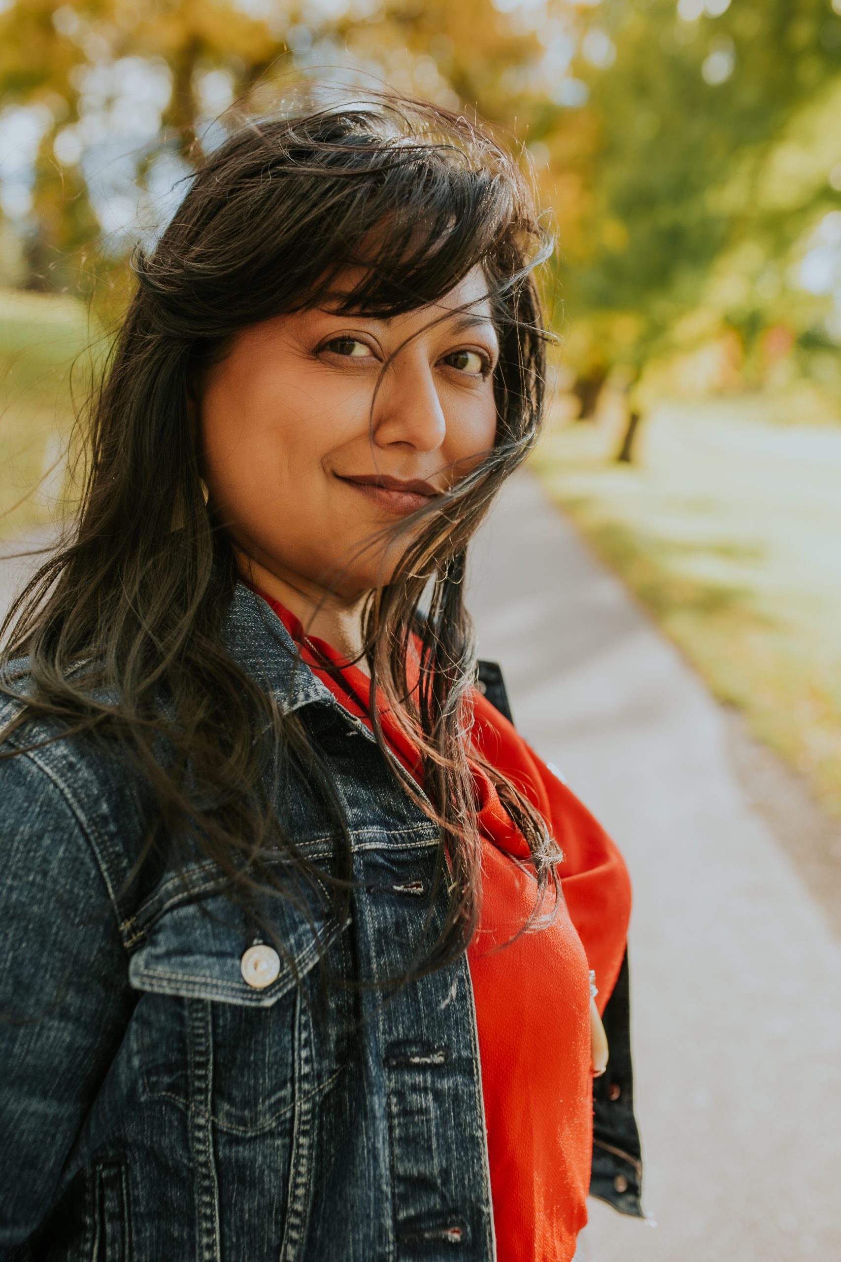 Portrait of Tyler in a denim jacket and red blouse on a tree-lined path