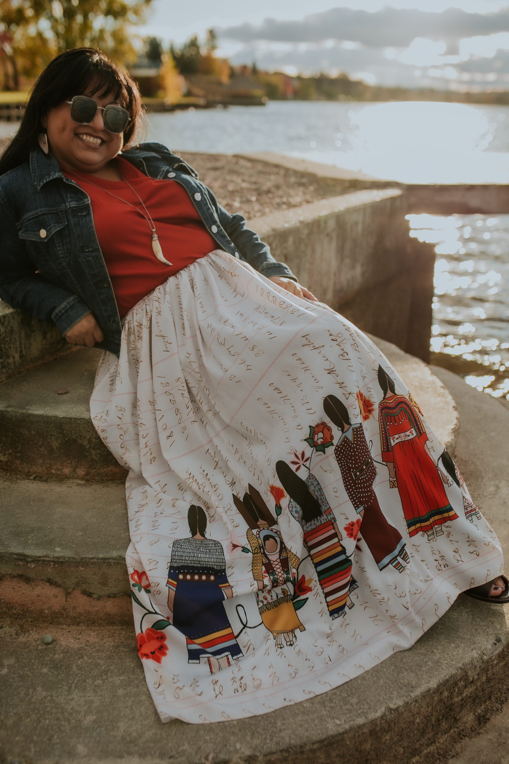 Tyler seated on a lakeside stone wall in a ribbon-art skirt, red blouse, and denim jacket