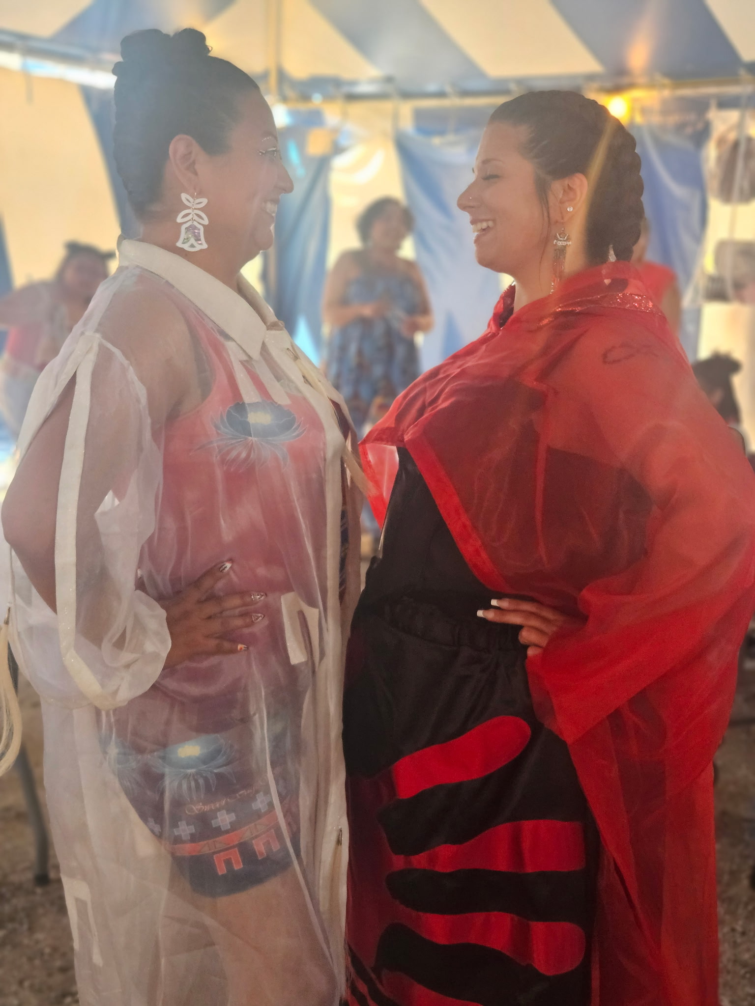 Two models in sheer red and white designer coats backstage at a festival tent